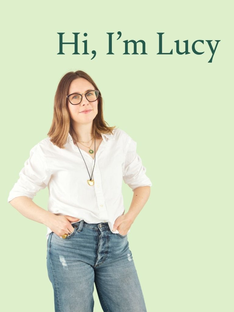 Hi, I'm Lucy. A white woman with brown hair, black rimmed glasses and a white shirt, smiles at the camera with her hands tucked into the front pockets of her jeans. 