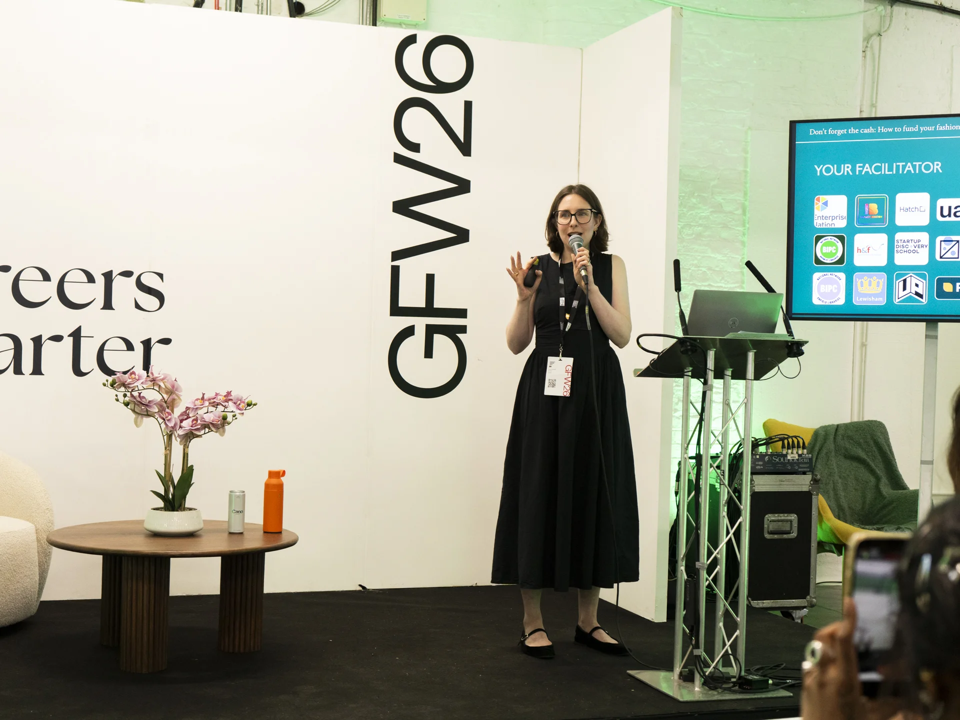 A white woman with short, brown hair wears black rimmed glasses and a black dress. She is standing on a stage with 'GFW26' printed behind her. She is holding a microphone and speaking to an audience.