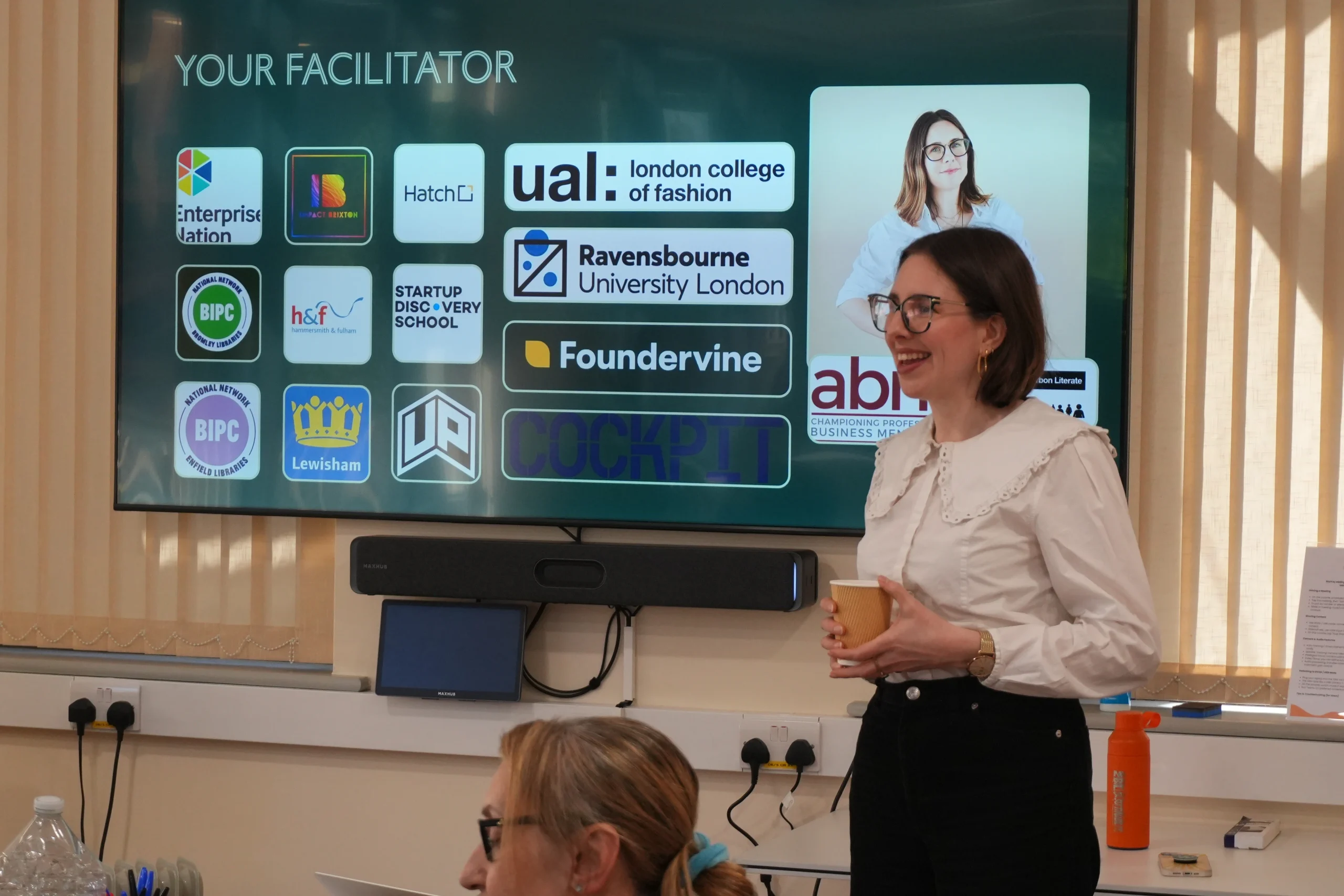 A white woman with short brown hair, wearing black rimmed glasses and a white shirt, stands in front of a presentation screen. She is smiling to someone off camera left and holding a paper cup. 