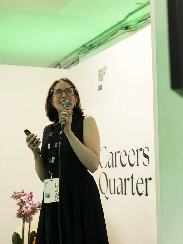 A portrait of a white woman wearing glasses and a black dress. She is smiling, holding a microphone in one hand and a clicker in her other. The text behind her reads 'GFW Careers Quarter'. 
